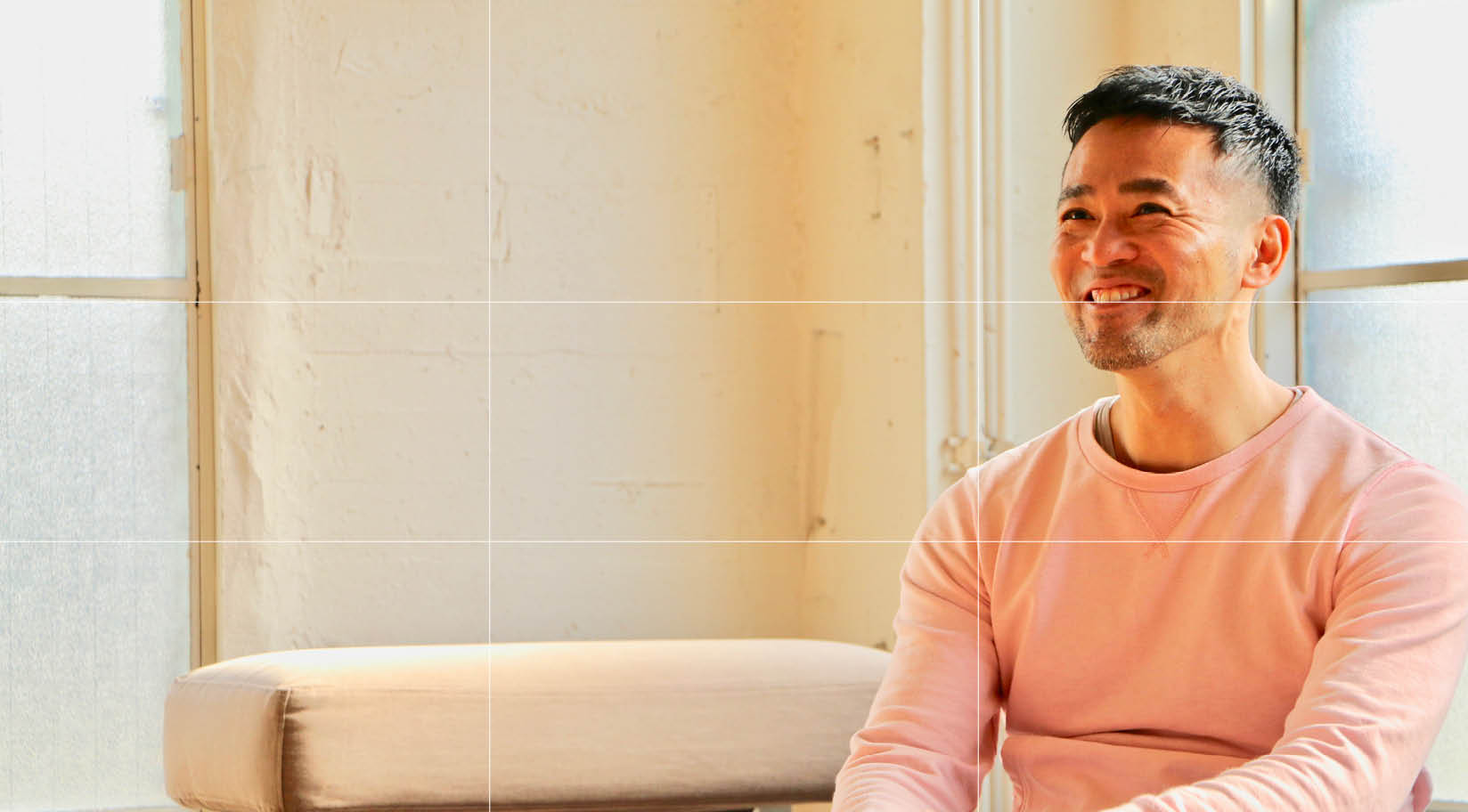 A young man, smiling warmly, positioned on the right side of the picture. He appears to be the interviewee, seated opposite the interviewer. The camera is at eye level to maintain a natural perspective. A grid overlay illustrates proper framing techniques, with plenty of light filling the room.