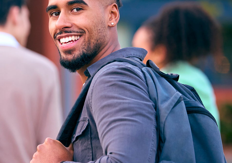 A student with a denim shirt and backpack looks over his shoulder and smiles at the camera