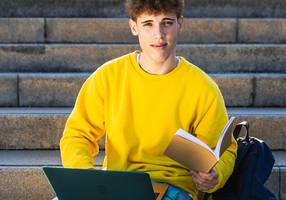 A student in a yellow jumper sits on some steps with a yellow notebook and a laptop