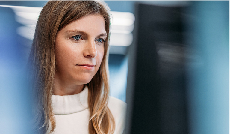 A woman with blonde hair and a white jumper is looking at a screen