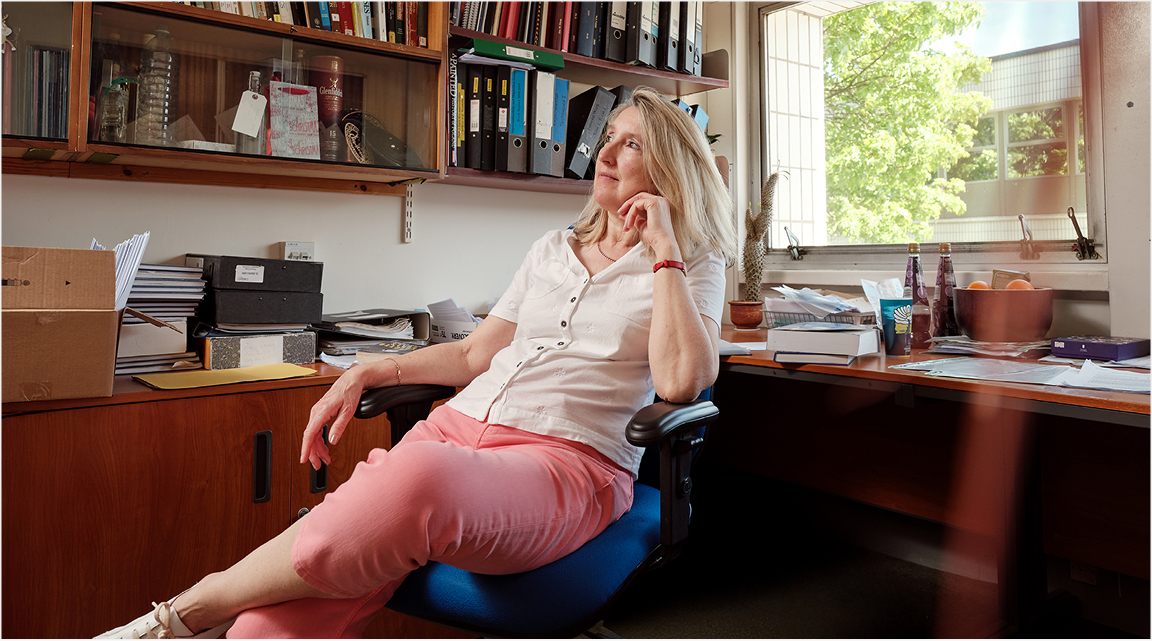 A woman in a beige shirt and coral trousers is sitting in her office, the shelves are lined with bookcases, she is staring into the distance as if deep in thought