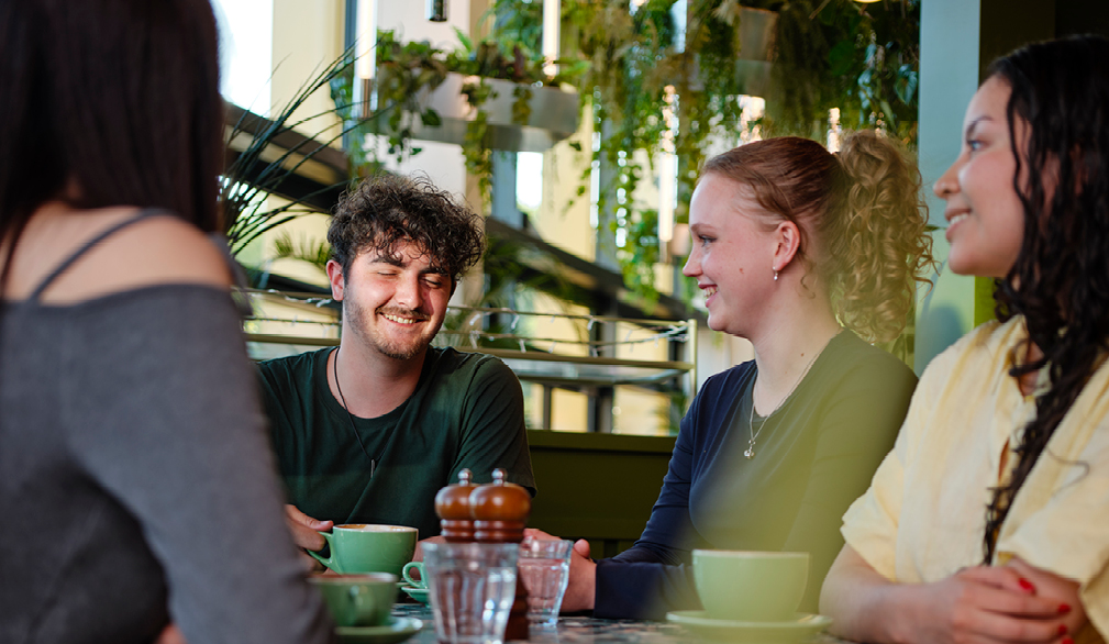 Four young people, three women and one man, sitting together in a bright, modern café. The space is filled with natural light and surrounded by greenery and potted plants. The group appears cheerful and engaged in conversation, smiling and relaxed.