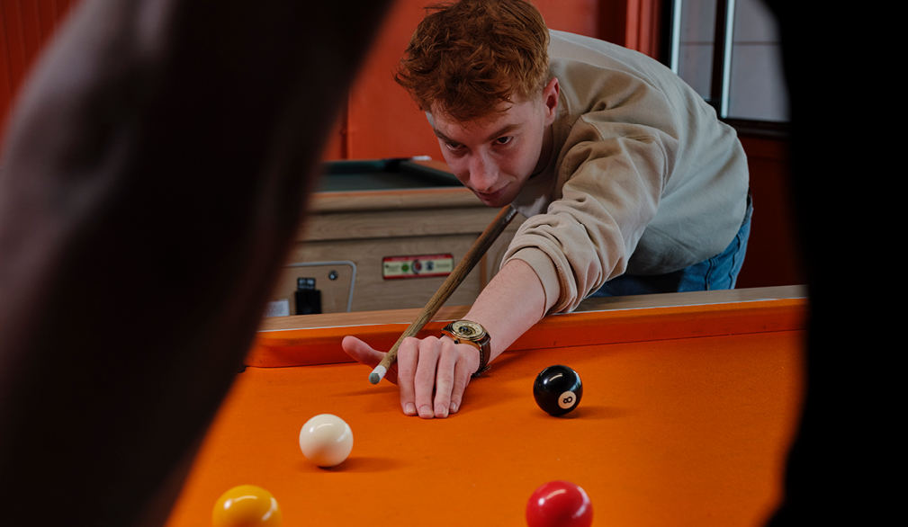 A young man holding a cue, preparing to take a shot at an orange snooker table. He is focused, leaning forward with the cue positioned, ready to play.