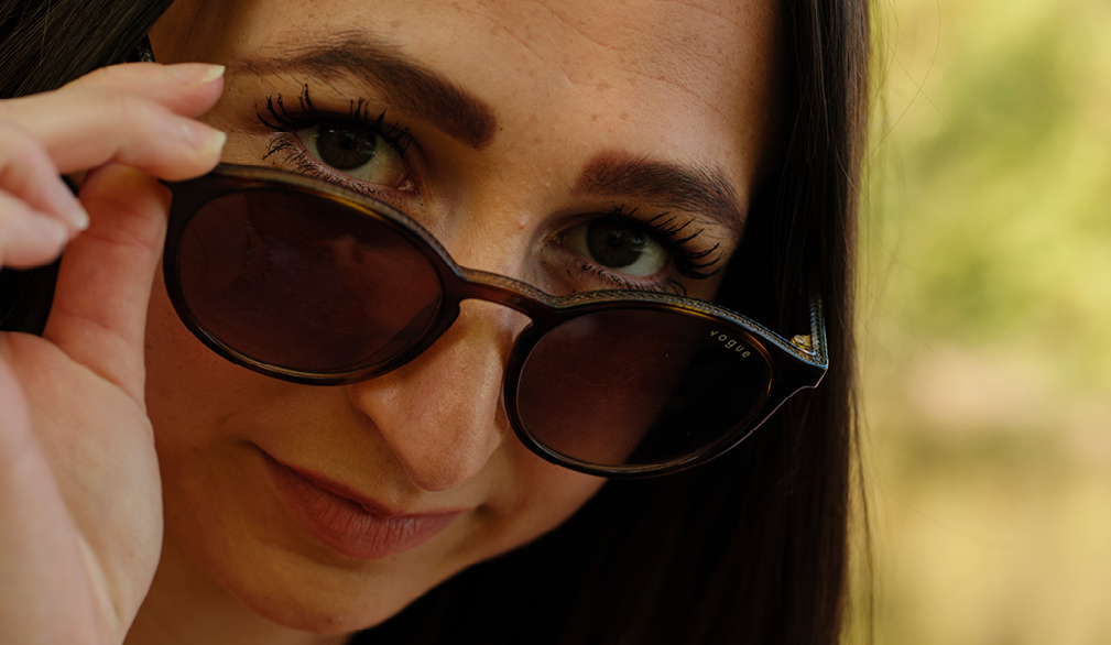 A woman looking directly at the camera while removing her sunglasses. The camera focuses on her face, and the sunglasses are halfway down her nose, as if she’s intentionally pausing mid-gesture.