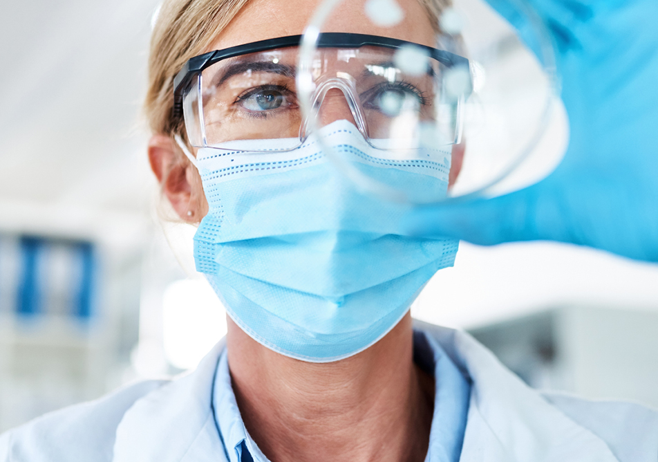 A scientist wearing goggles and a mask is looking at the contents of a petri dish