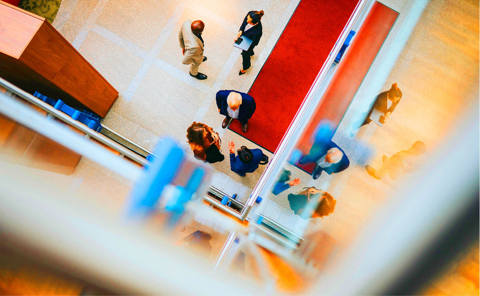 An oversaturated photo taken indoors from above. Below, there are two groups of people talking, all dressed smartly. In one group, a woman holds a laptop while a man stands with his hand behind his back. The other group has a woman and two men, with one man gesturing as the others listen. Their reflections can be seen on nearby glass surfaces.