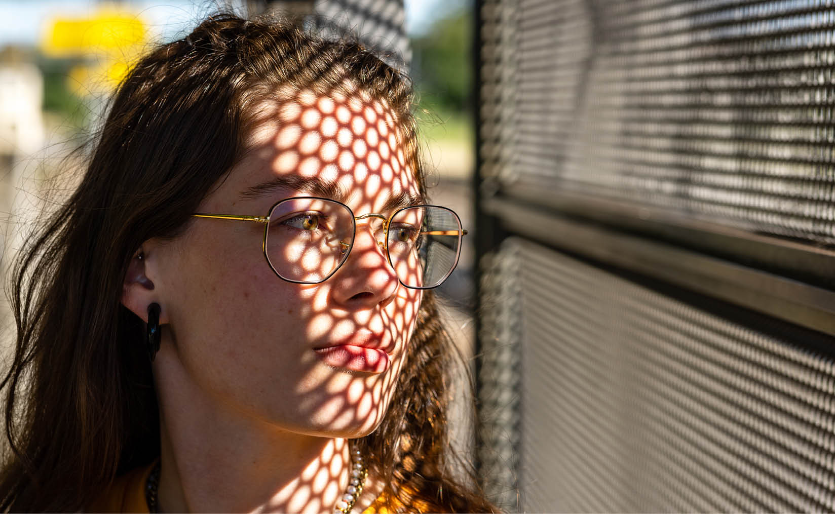 A photo of a young girl outdoors, positioned on the left side of the picture, looking to the right through a wall with many small honeycomb-shaped holes. Light filters softly through the holes, partially illuminating her face. She wears a thoughtful, dreamy expression.