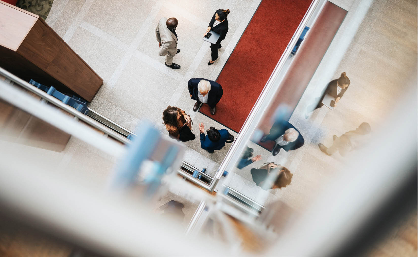 A photo taken indoors from above. Below, there are two groups of people talking, all dressed smartly. In one group, a woman holds a laptop while a man stands with his hand behind his back. The other group has a woman and two men, with one man gesturing as the others listen. Their reflections can be seen on nearby glass surfaces.