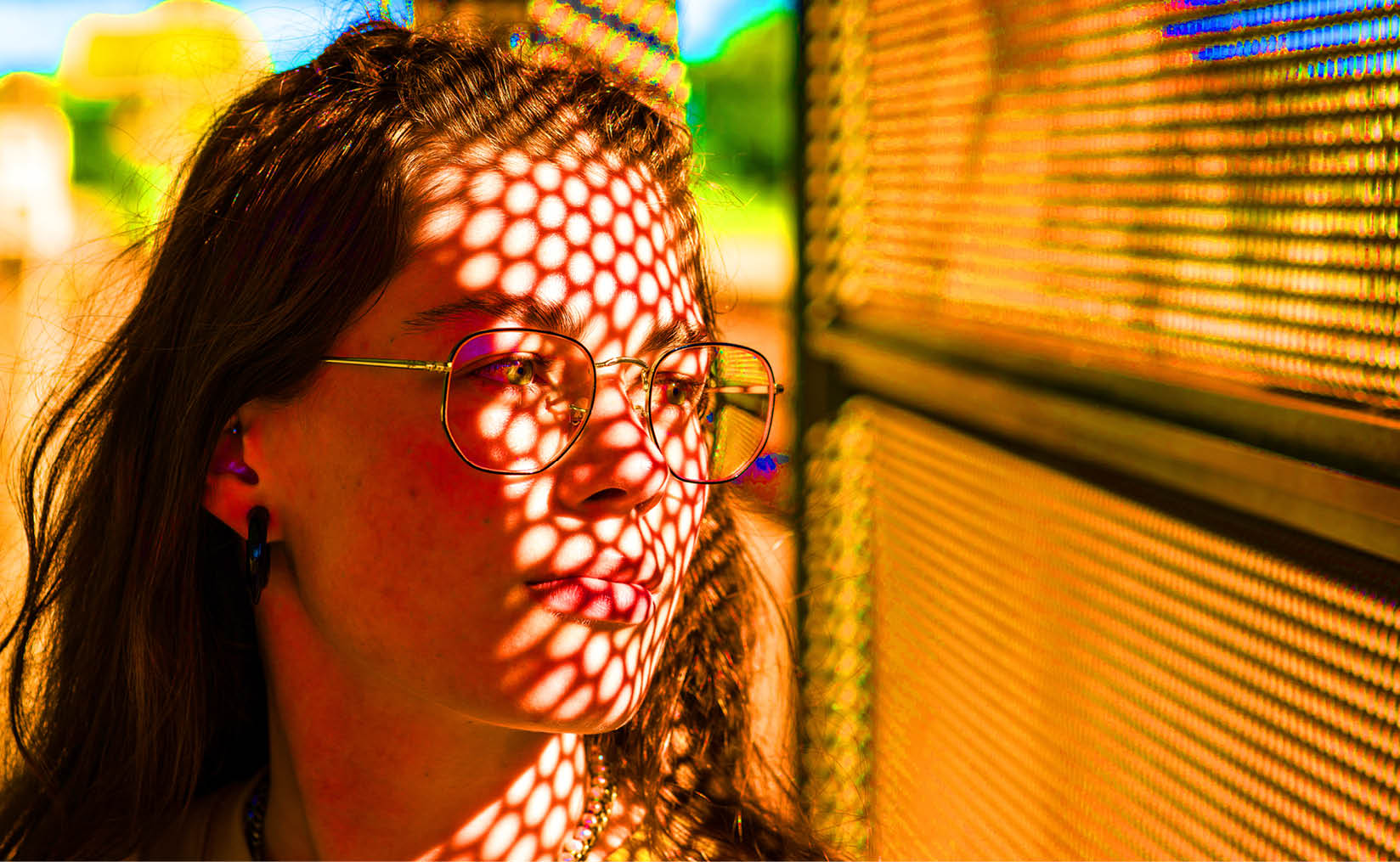 An oversaturated photo of a young girl outdoors, positioned on the left side of the picture, looking to the right through a wall with many small honeycomb-shaped holes. Light filters softly through the holes, partially illuminating her face. She wears a thoughtful, dreamy expression.
