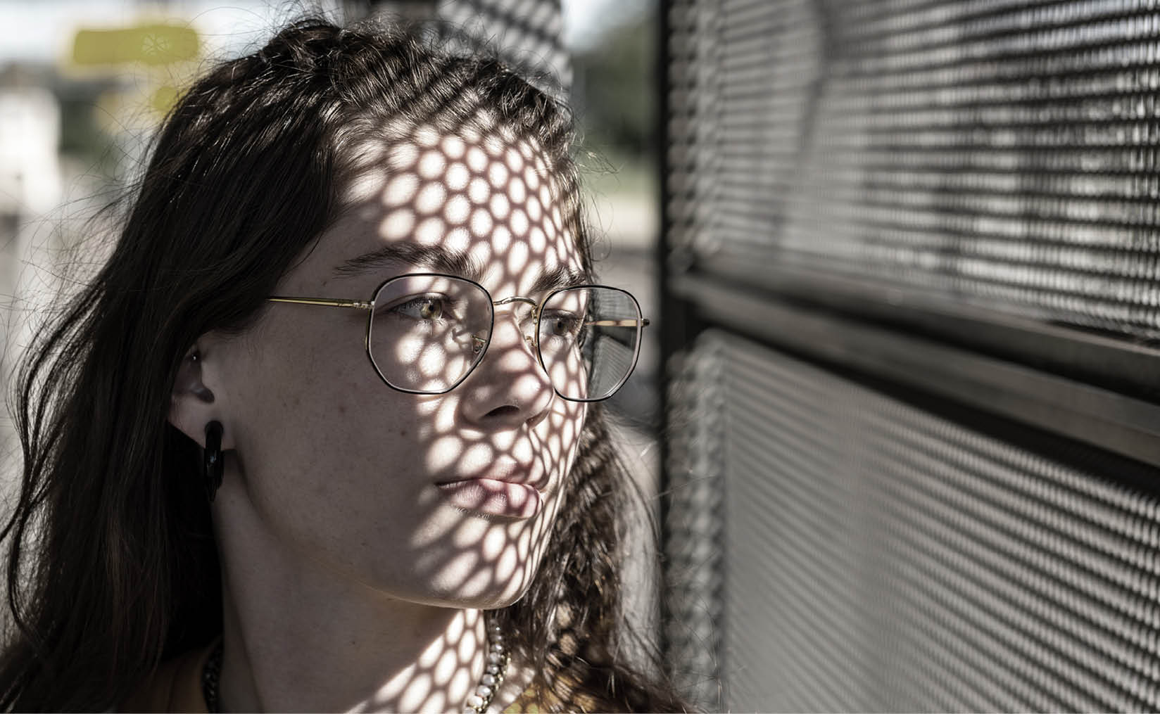 An under-saturated photo of a young girl outdoors, positioned on the left side of the picture, looking to the right through a wall with many small honeycomb-shaped holes. Light filters softly through the holes, partially illuminating her face. She wears a thoughtful, dreamy expression.