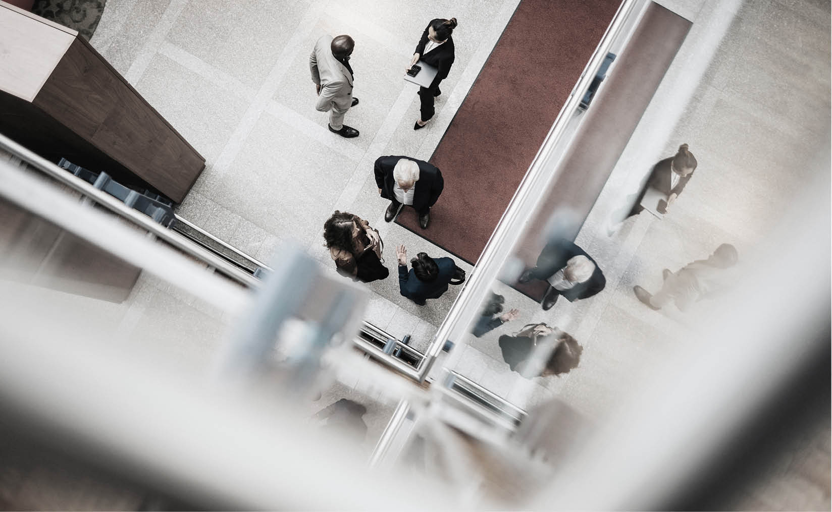 An under-saturated photo taken indoors from above. Below, there are two groups of people talking, all dressed smartly. In one group, a woman holds a laptop while a man stands with his hand behind his back. The other group has a woman and two men, with one man gesturing as the others listen. Their reflections can be seen on nearby glass surfaces.