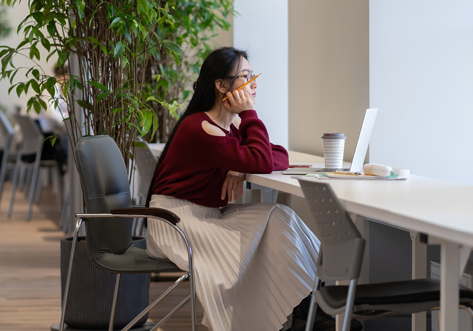 A student sits at a desk looking thoughtful and holding a pencil, they have a laptop and a reusable coffee cup in front of them