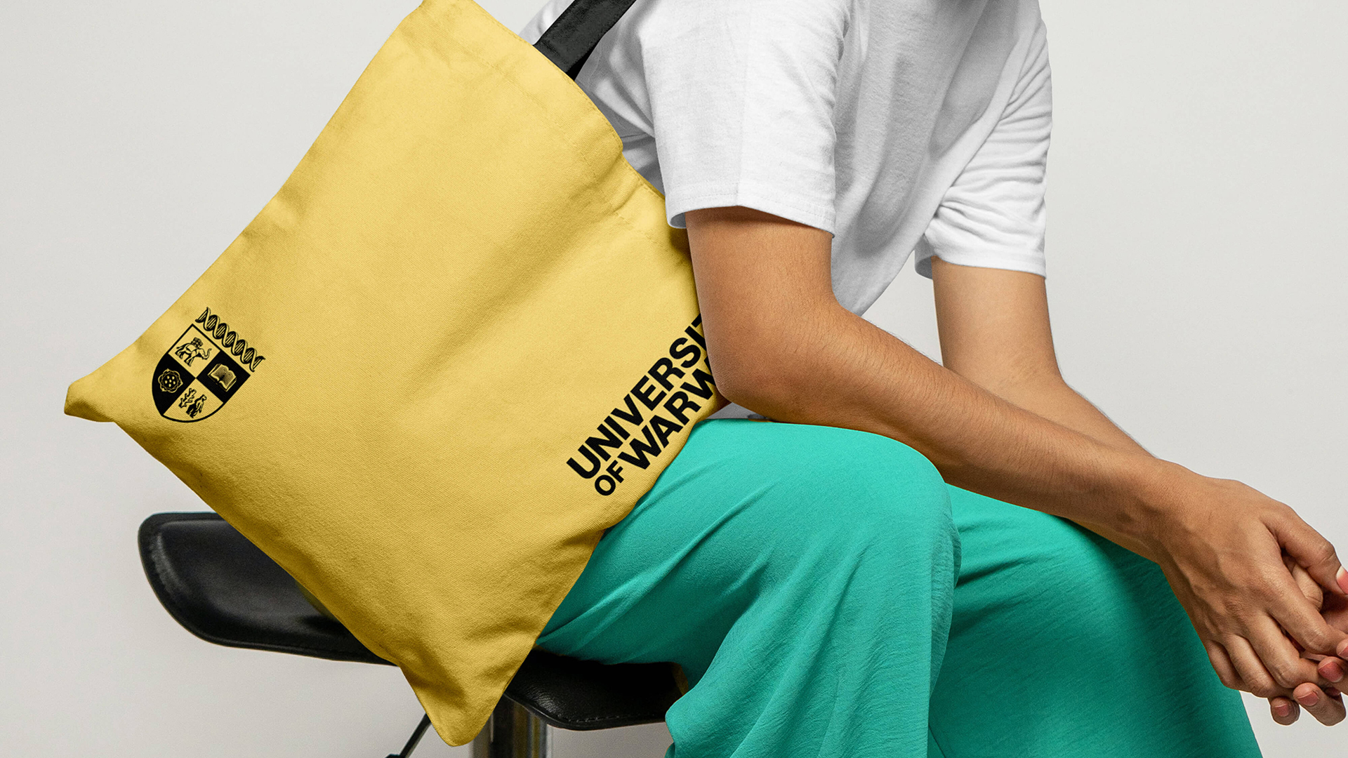 A person wearing a white t-shirt and green trousers sits on a stool while holding a yellow University of Warwick-branded canvas bag over their shoulder.