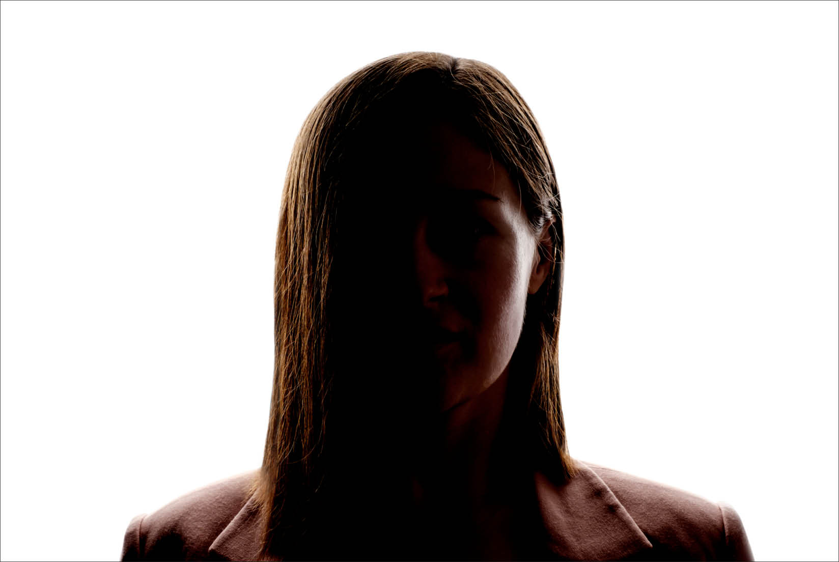 A young woman with a neutral expression looking directly at the camera. She’s centered in the frame, visible from the neck up, but her features are obscured due to high-contrast lighting.