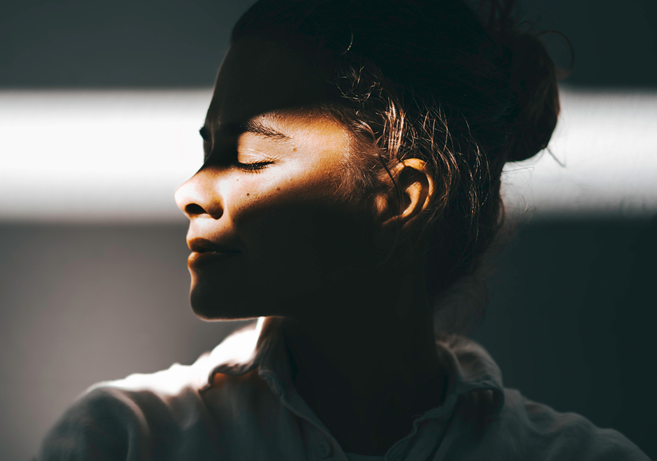 A woman looking left with her eyes closed, the room is dark but a strip of light illuminates her face