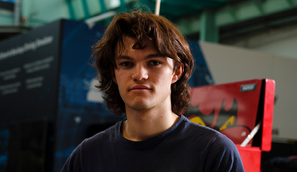 A young man indoors, centered in the frame, looking directly at the camera with a neutral expression.