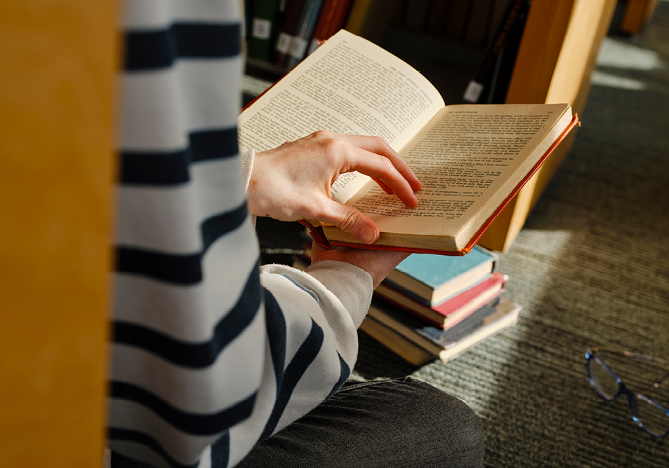 A person is reading a book while sat against a shelf viewed as if sitting next to them