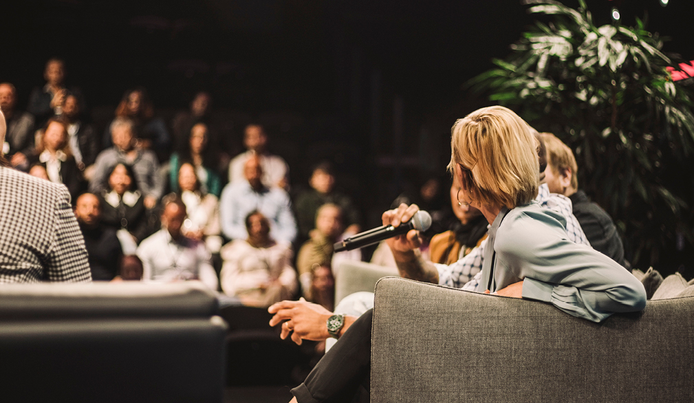 A group of people seated on a stage in an amphitheatre setting, with their backs to the camera. One woman is speaking into a microphone, while some of the other speakers turn to look at her as she addresses the audience.
