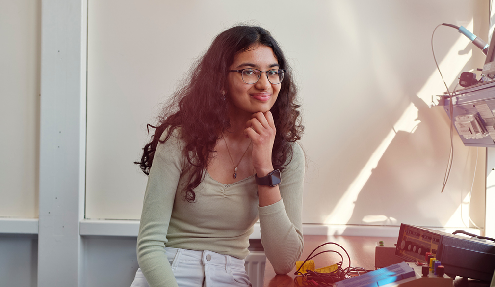 A young woman sitting by a classroom window with sunlight streaming in. She’s smiling gently, resting her chin on one hand, while her other hand rests on her tights.