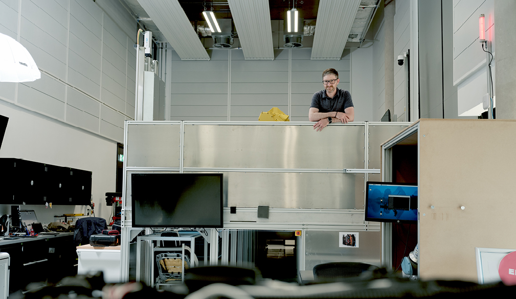 A middle-aged man with a gentle smile looking toward the camera from a distance. He stands on a platform with his lower body hidden behind a wall, inside a room with electronic equipment.