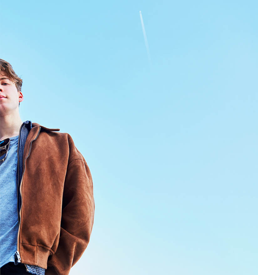 Graded footage showing the upper half of a young boy, cropped to one side, standing against a bright blue sky. He’s looking down at the camera below, with a faint white trail from a distant airplane visible in the sky.